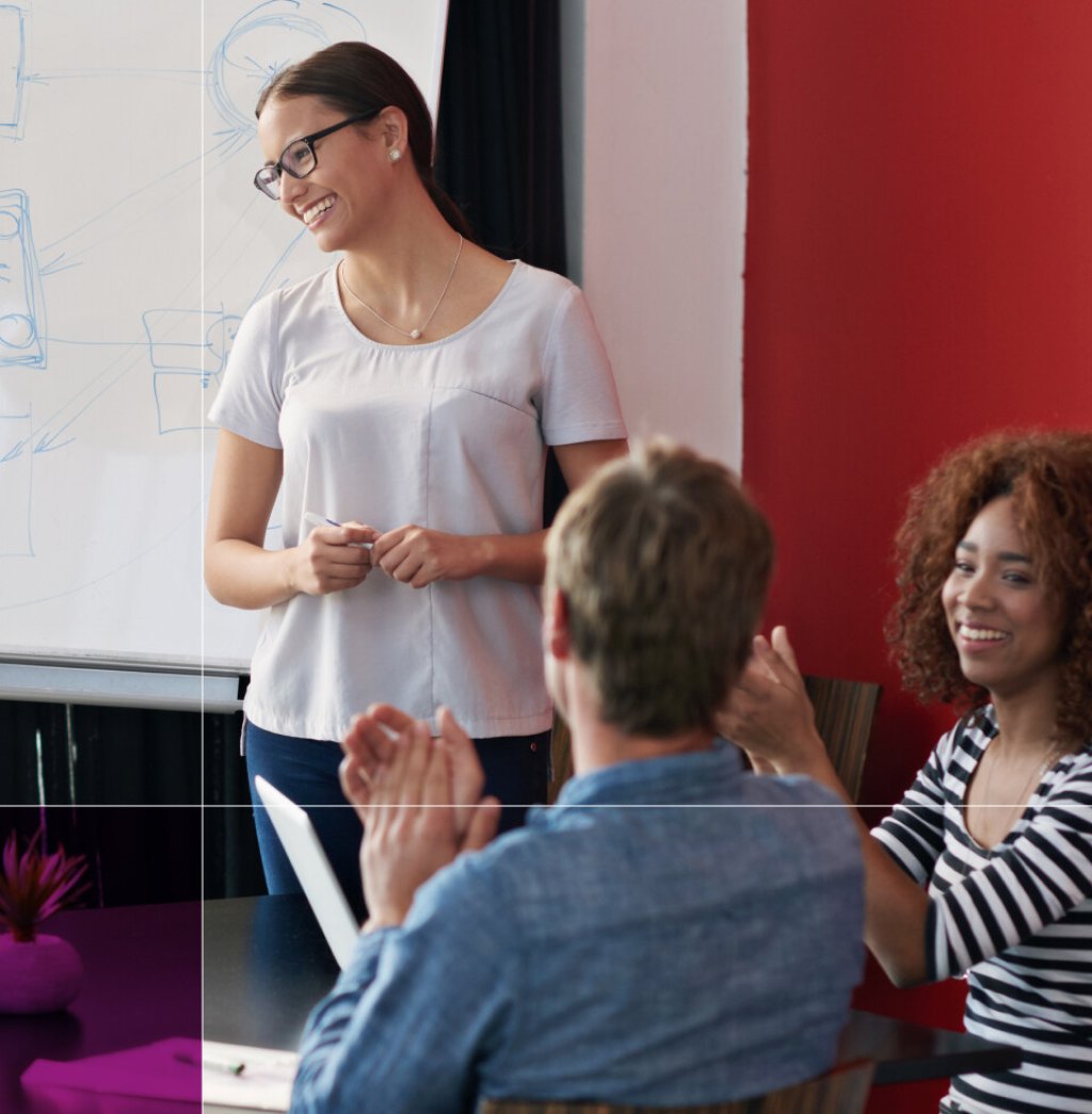 Woman presenting at an event. She is at a white board and two members of the audience can be seen applauding her.